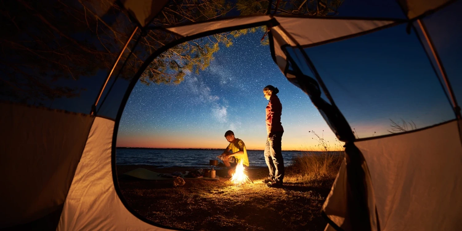 View from the inside of a tent looking out. Woman standing next to a campfire, man sitting, and lake at the background