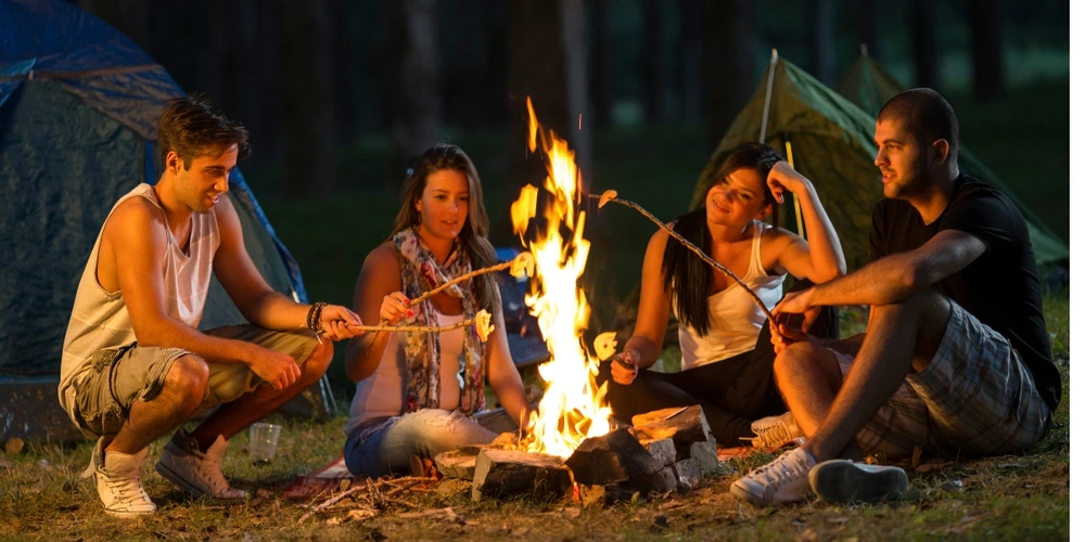 Four people, 2 male and 2 female, camping by the fire and toasting marshmallows