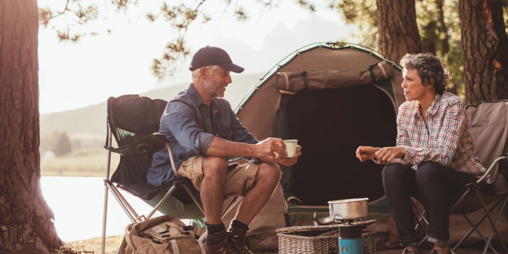 Happy mature campers enjoying coffee by the lake in front of their tent
