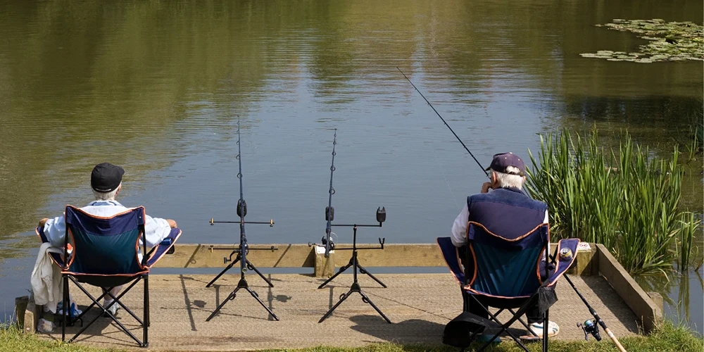 Two elderly men who enjoy fishing sitting at the edge of the lake waiting for the fish to bite